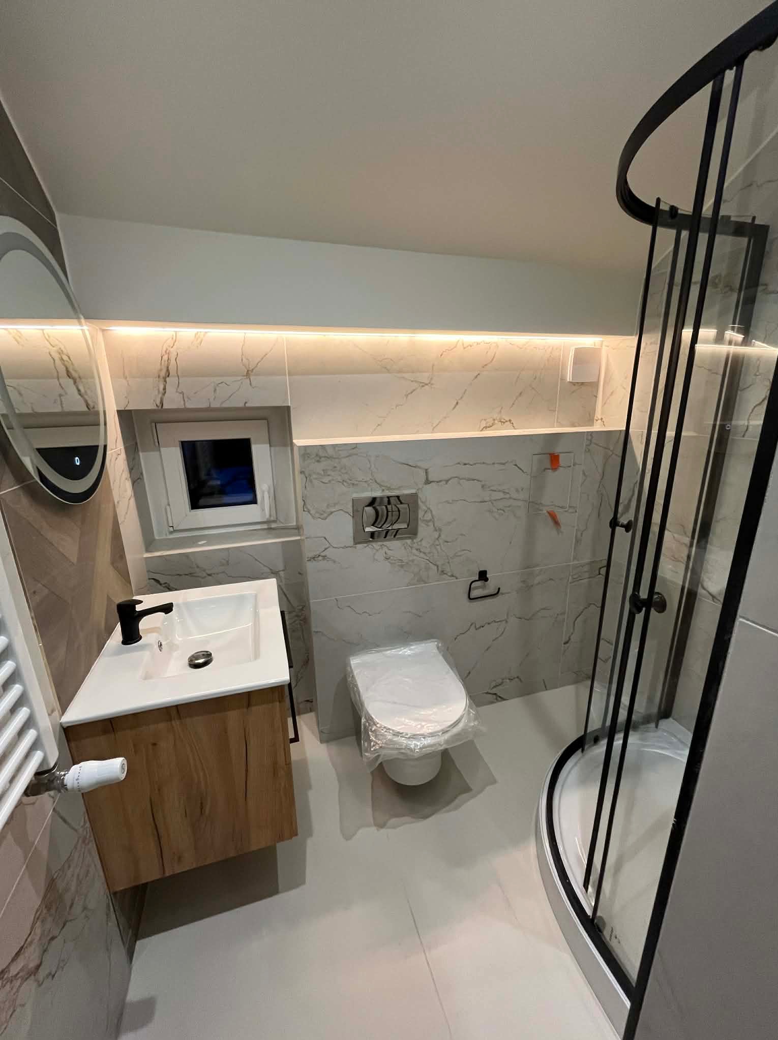 Nowoczesna łazienka po remoncie: kabina prysznicowa z czarnymi ramami, umywalka z drewnianą szafką, marmurowe płytki i podświetlane lustro. Toaleta zabezpieczona folią.