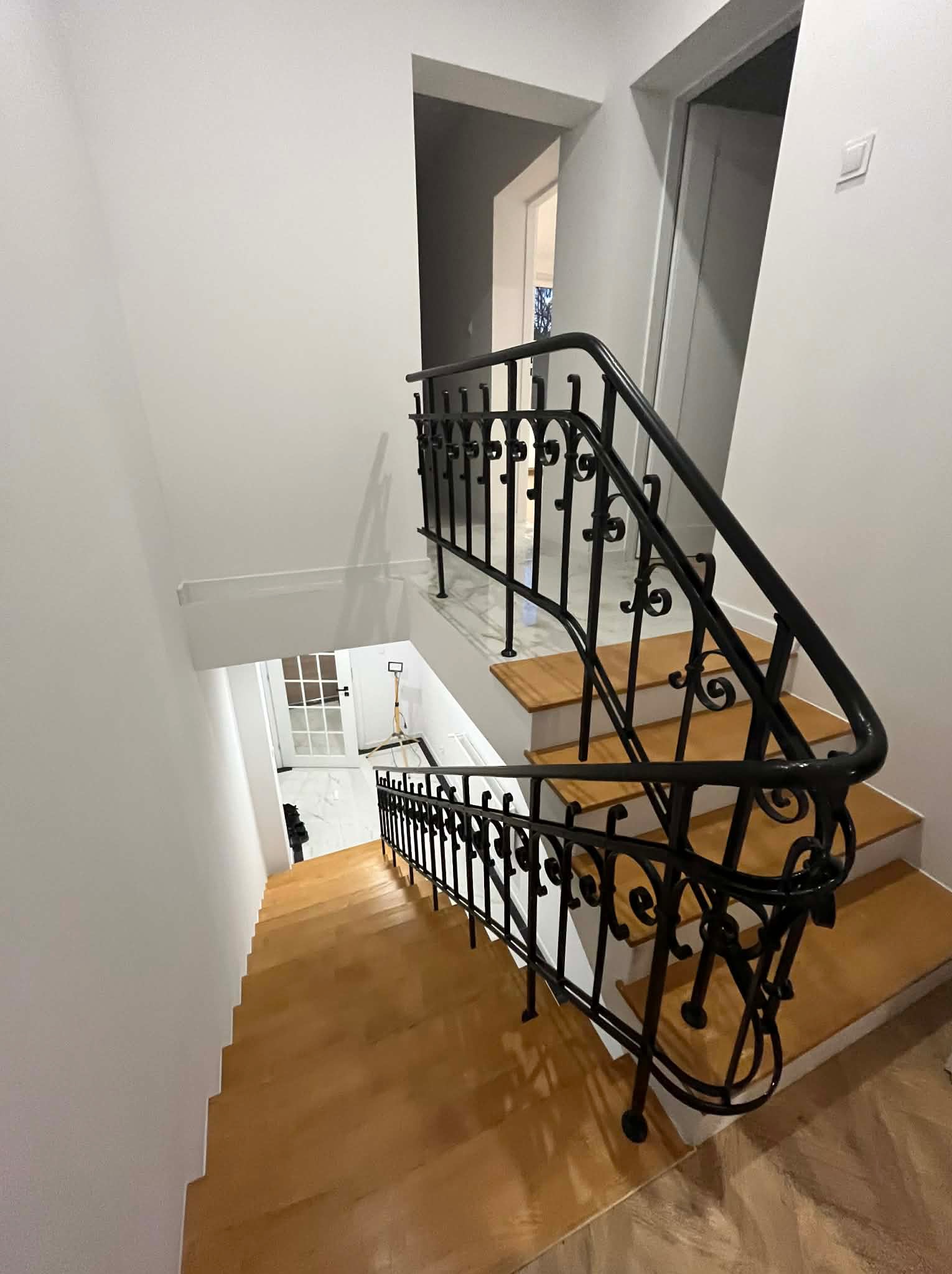 Klatka schodowa z drewnianymi stopniami i ozdobną, czarną, kutą balustradą. Widok z góry na dół, białe ściany, drzwi w tle. Charakterystyczny wzór balustrady.