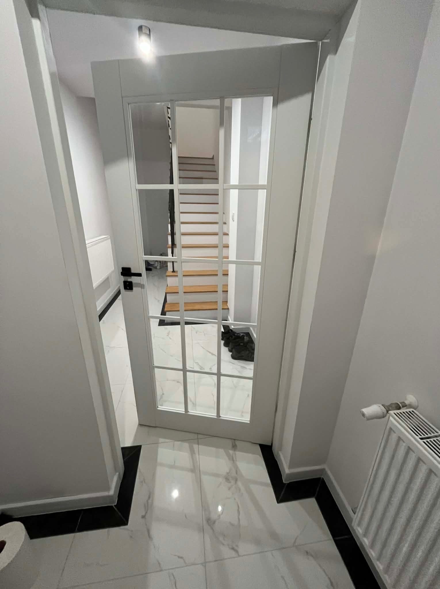 Białe, przeszklone drzwi wewnętrzne z czarnymi klamkami, widok na schody w jasnym wnętrzu z marmurową podłogą i grzejnikiem. Minimalistyczny design.