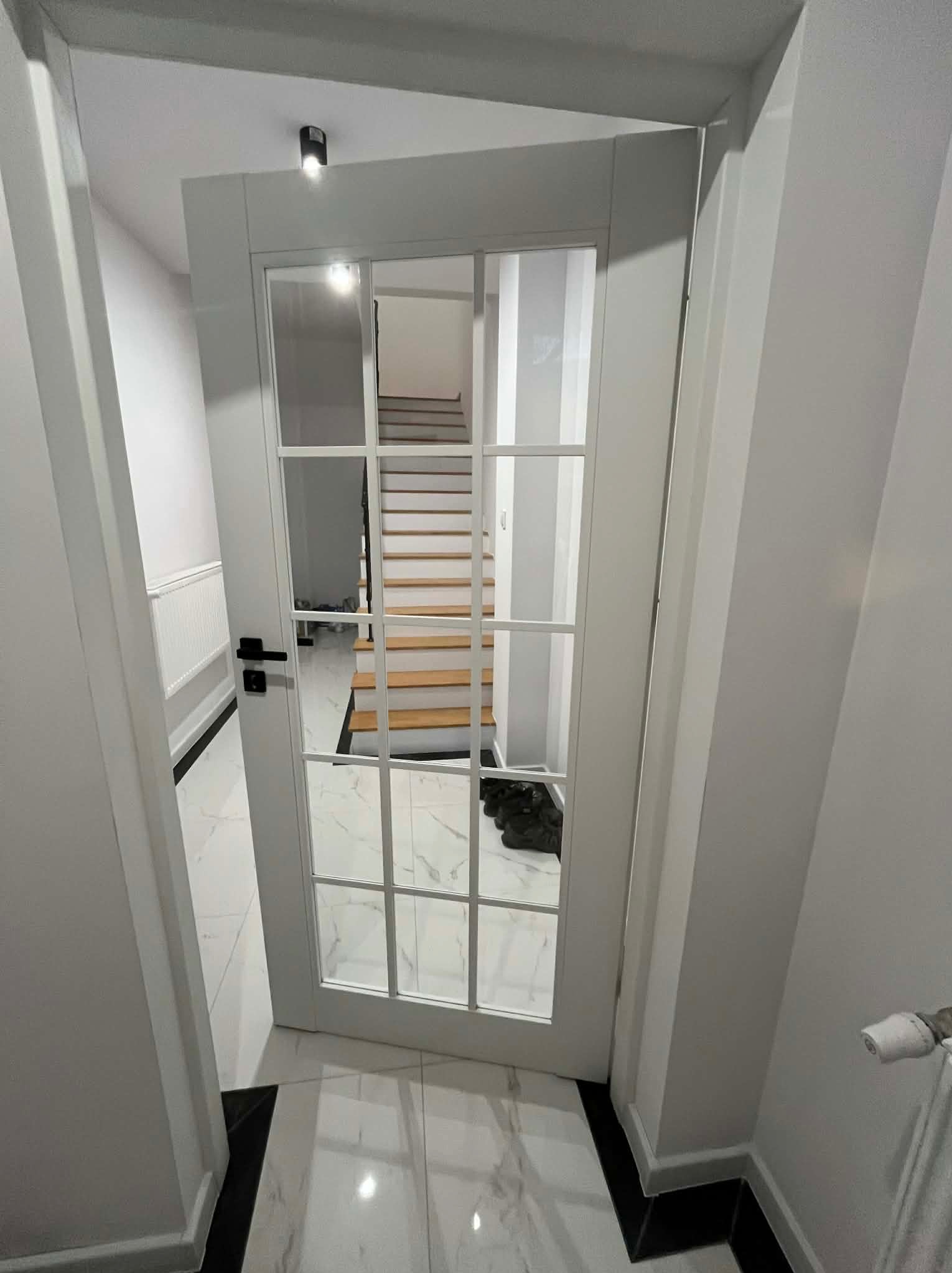 Białe, przeszklone drzwi wewnętrzne z czarną klamką, widok na schody i marmurową podłogę. Minimalistyczny design, jasne wnętrze.