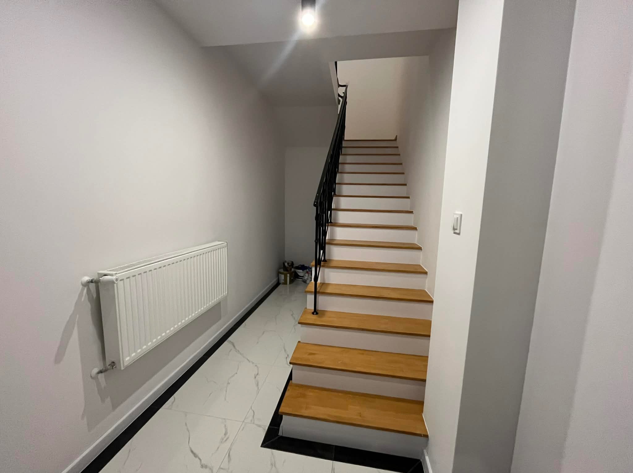 Białe wnętrze z drewnianymi schodami i grzejnikiem na ścianie. Marmurowe płytki podłogowe z czarnymi akcentami. Minimalistyczny design, oświetlenie sufitowe.