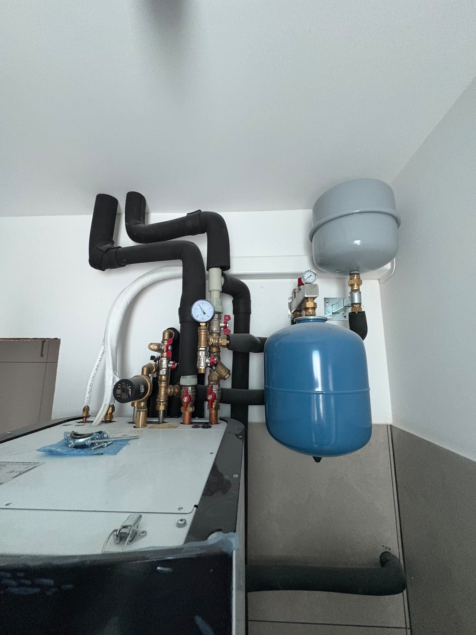 Złożona instalacja grzewcza z czarnymi rurami, manometrem i niebieskim zbiornikiem wyrównawczym. Widoczne elementy montażowe i białe ściany w tle.
