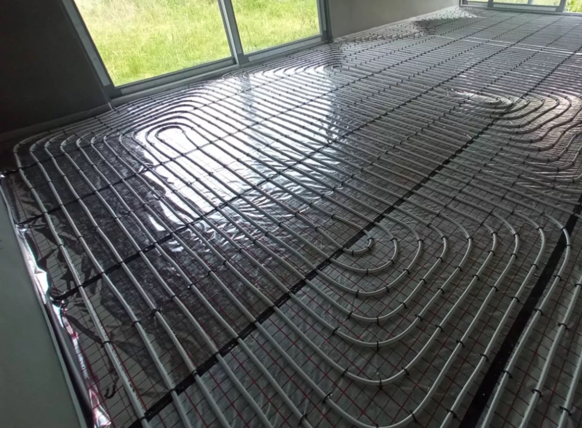 Białe rury ogrzewania podłogowego ułożone na folii izolacyjnej z siatką, widok z góry, duże okno w tle. Etap instalacji przed wylewką.