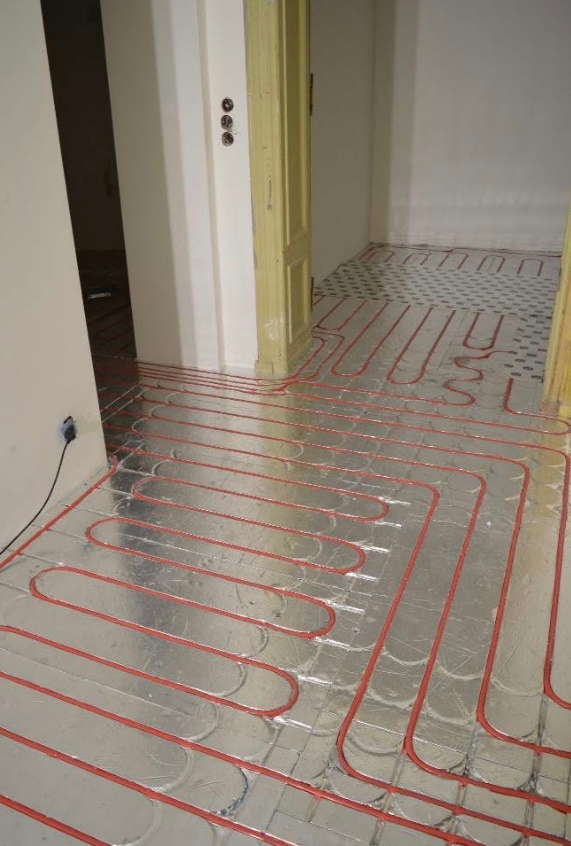 Czerwone rury ogrzewania podłogowego ułożone na srebrnej folii w jasnym wnętrzu, widoczne fragmenty podłogi z wzorem i otwarte drzwi do innego pomieszczenia.
