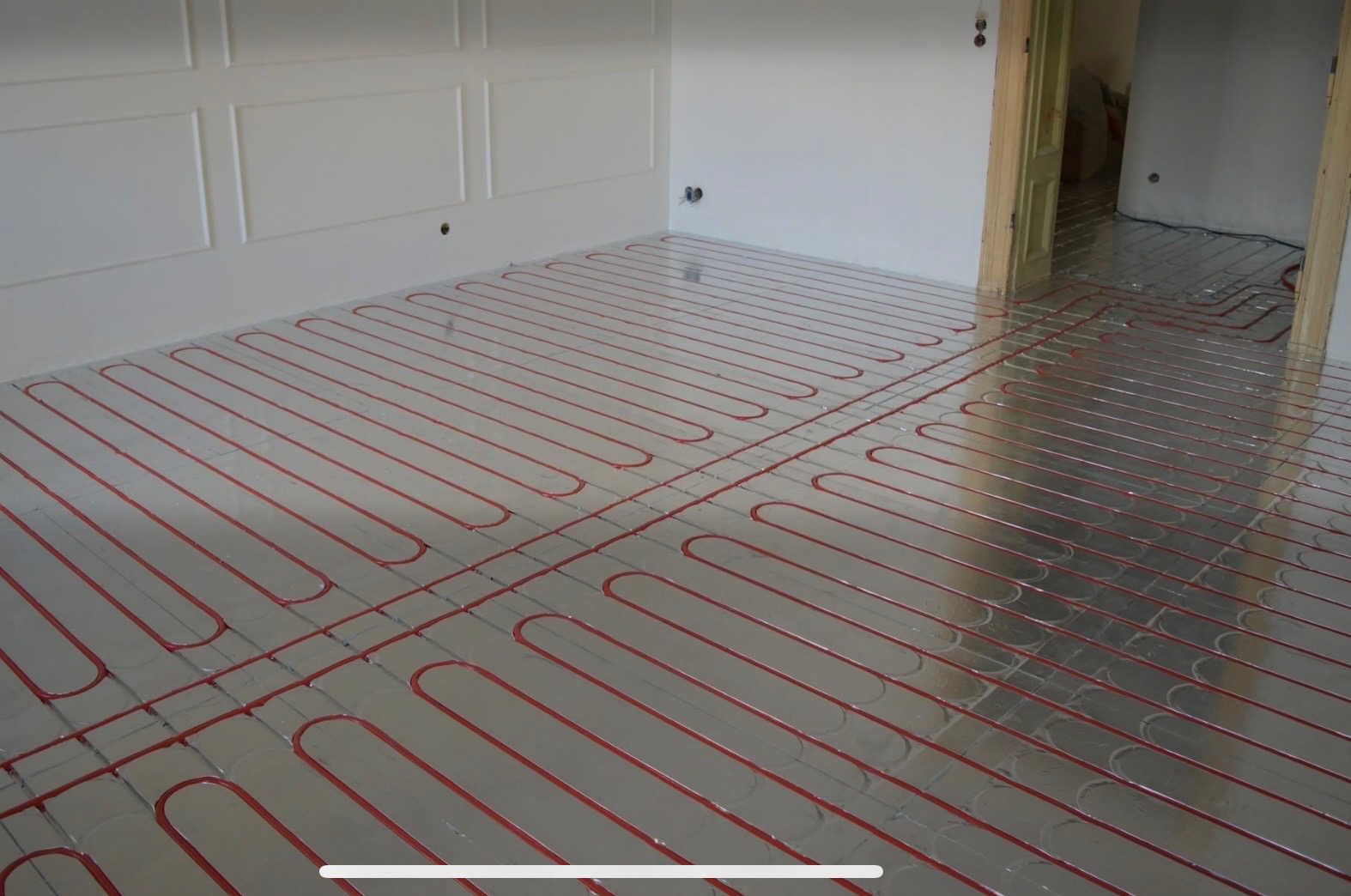 Ułożone, czerwone rury ogrzewania podłogowego na srebrnej folii izolacyjnej w jasnym pomieszczeniu z dekoracyjną ścianą i otwartymi drzwiami w tle.