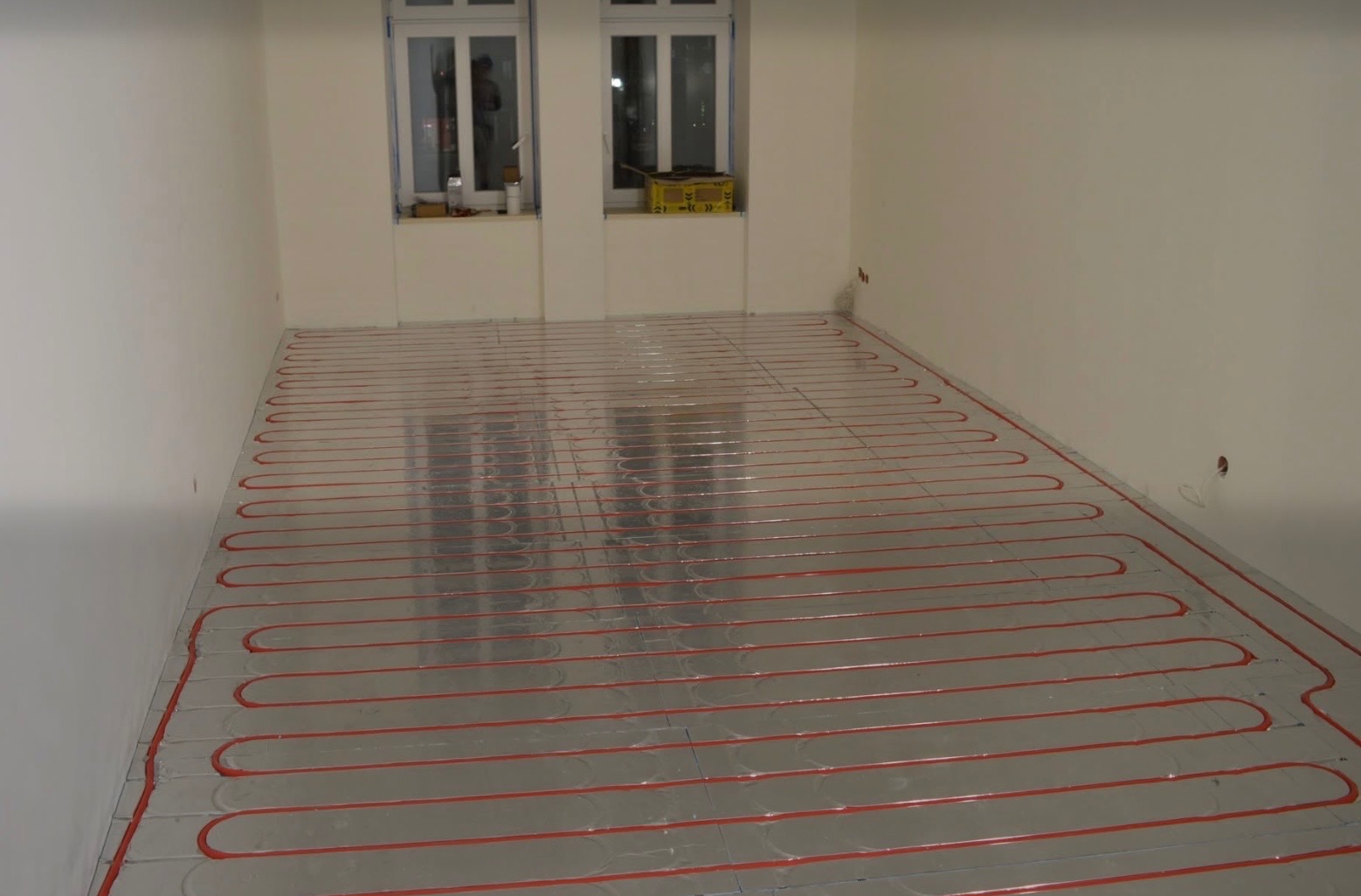 Ułożona instalacja ogrzewania podłogowego w pustym, jasnym pomieszczeniu z dwoma oknami. Czerwone rurki tworzą pętle na srebrnej folii izolacyjnej.