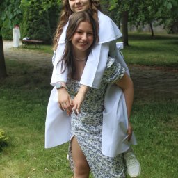 Uwaga Flash - Uśmiechnięta nastolatka w kwiecistej sukience daje barana dziewczynce ubranej w białą albę komunijną i wianek na głowie, na tle zielonego trawnika i drzew.