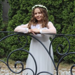 Uwaga Flash - Uśmiechnięta dziewczynka w białej sukience komunijnej i wianku na głowie, oparta o ozdobną, metalową barierkę na tle zielonych krzewów.