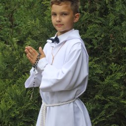 Uwaga Flash - Chłopiec w białej albie, z różańcem w dłoniach, pozuje na tle zielonych krzewów.