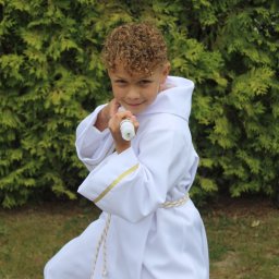 Uwaga Flash - Uśmiechnięty chłopiec w białej albie komunijnej, z kręconymi włosami, trzymający świecę na tle zielonego żywopłotu.