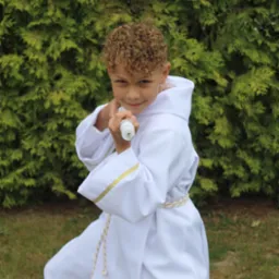 Uśmiechnięty chłopiec w białej albie komunijnej, z kręconymi włosami, trzymający świecę na tle zielonego żywopłotu.