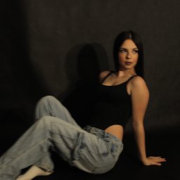 Uwaga Flash - Młoda kobieta w czarnym body i luźnych jeansach siedzi na podłodze na tle ciemnego tła, patrząc w bok.