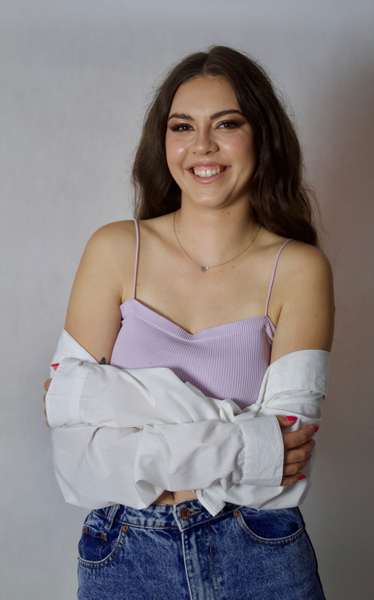 Uśmiechnięta, młoda kobieta z długimi brązowymi włosami, ubrana w liliowy top na ramiączkach, białą koszulę i jeansy, pozuje na jasnym tle.