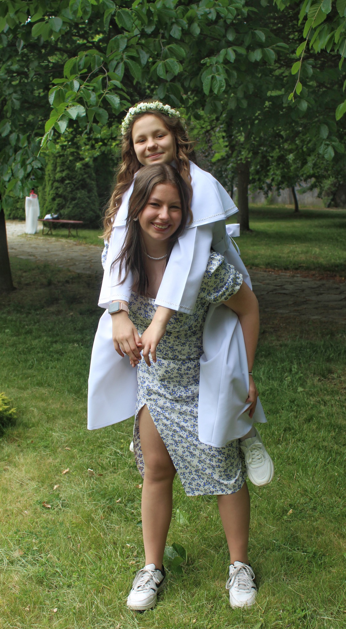 Uśmiechnięta nastolatka w kwiecistej sukience daje barana dziewczynce ubranej w białą albę komunijną i wianek na głowie, na tle zielonego trawnika i drzew.