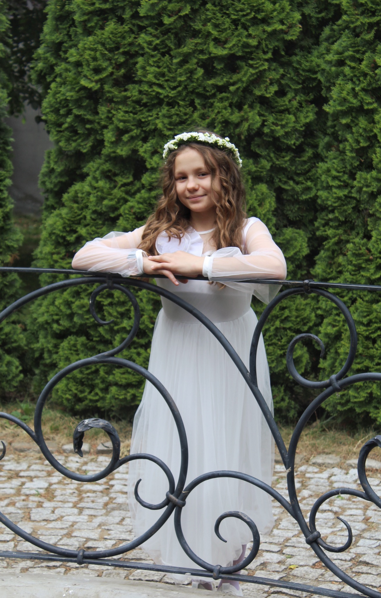 Uśmiechnięta dziewczynka w białej sukience komunijnej i wianku na głowie, oparta o ozdobną, metalową barierkę na tle zielonych krzewów.