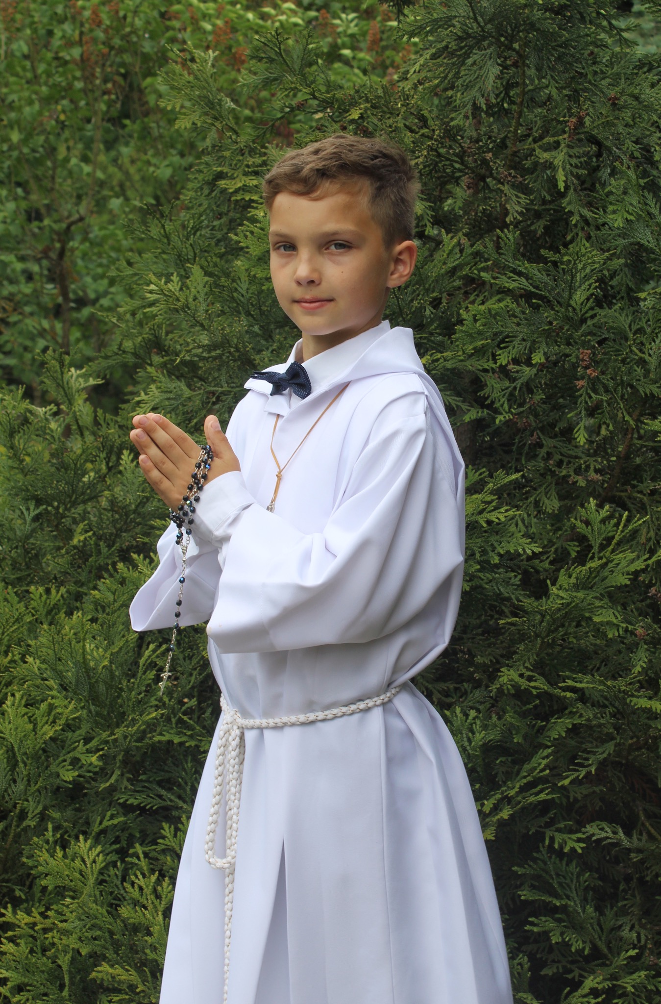 Chłopiec w białej albie, z różańcem w dłoniach, pozuje na tle zielonych krzewów.