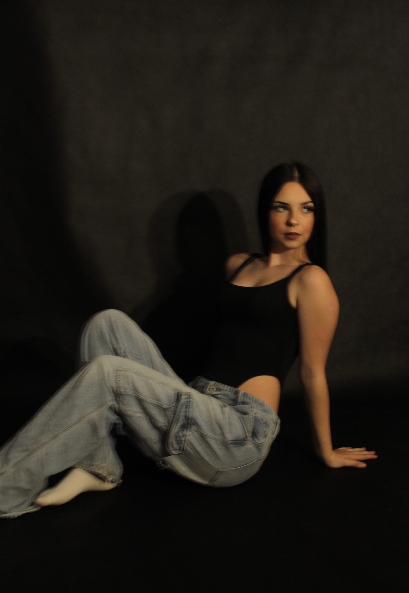 Młoda kobieta w czarnym body i luźnych jeansach siedzi na podłodze na tle ciemnego tła, patrząc w bok.