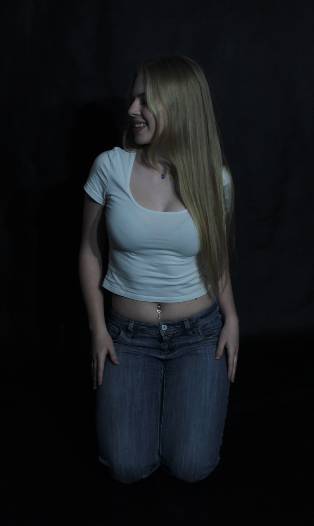 Portret kobiety na kolanach w białej koszulce i jeansach, z piercingiem w pępku i długimi blond włosami, uśmiechająca się w lewo na ciemnym tle.