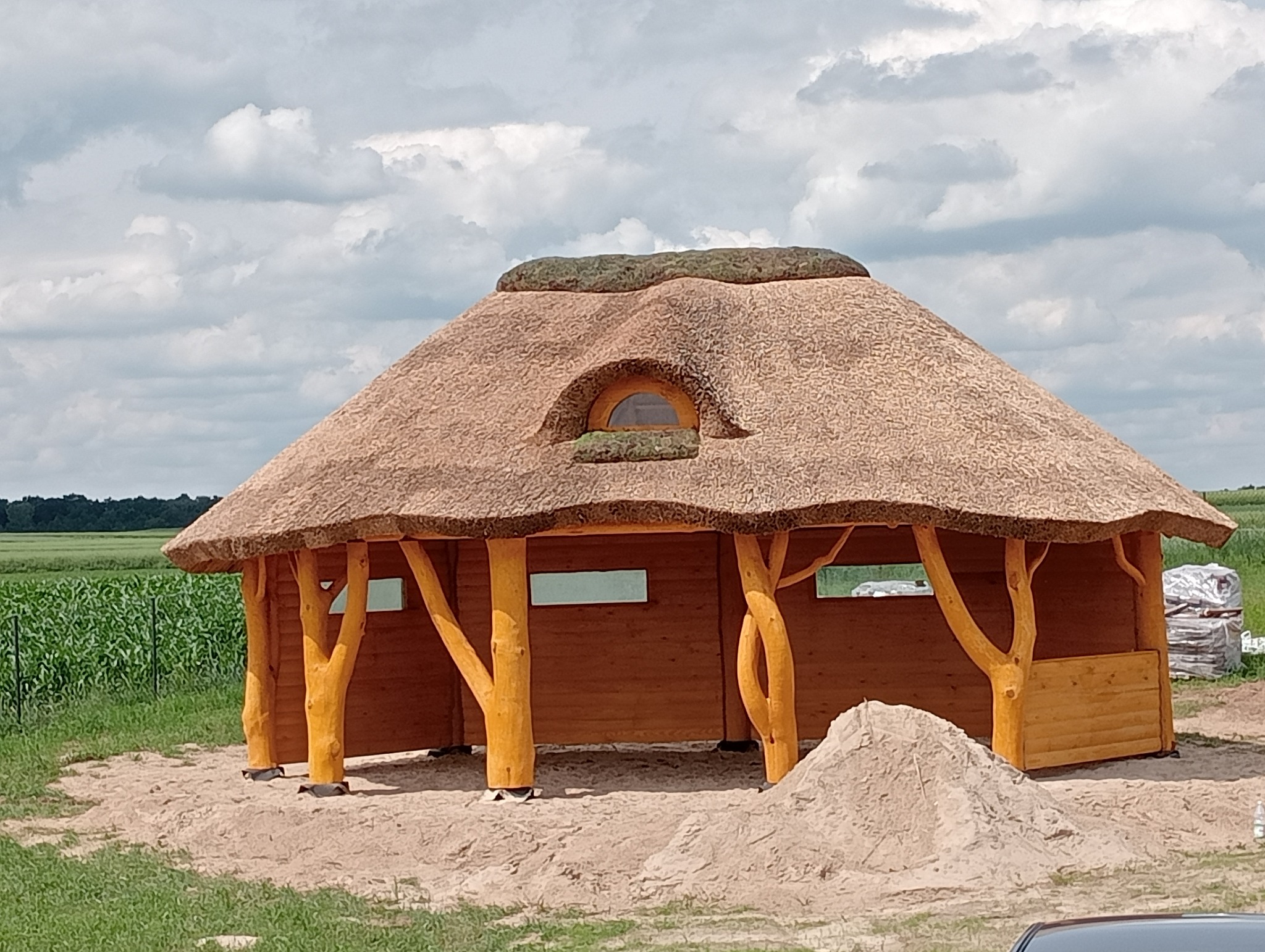 Drewniana altana z krytym strzechą dachem i małym okrągłym okienkiem, wsparta na pniach drzew, w tle pole kukurydzy i pochmurne niebo.