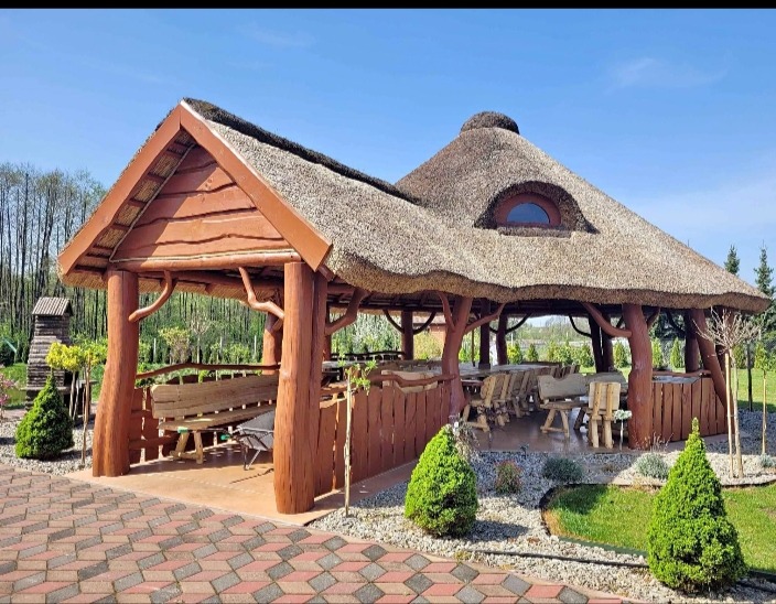 Kryta strzechą altana z drewnianymi filarami i meblami ogrodowymi, widok z zewnątrz na tle zieleni i błękitnego nieba.