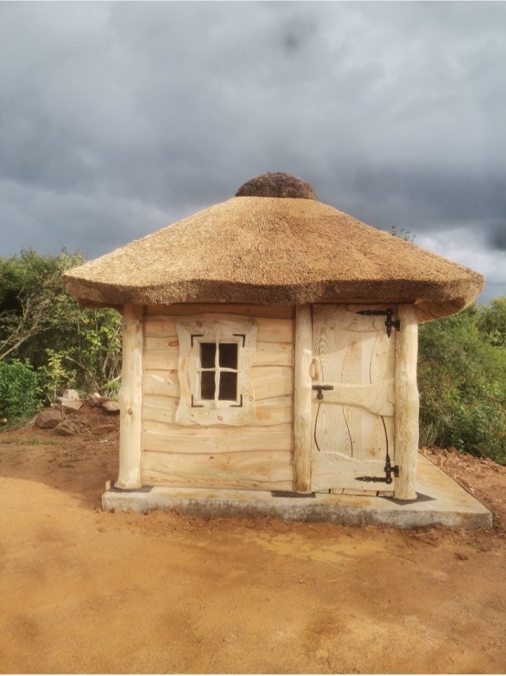 Mała, kwadratowa altana z bali z krytym strzechą dachem, umieszczona na betonowej podstawie, z widocznym oknem z ozdobną ramą i drzwiami z kutymi zawiasami na tle pochmurnego nieba.