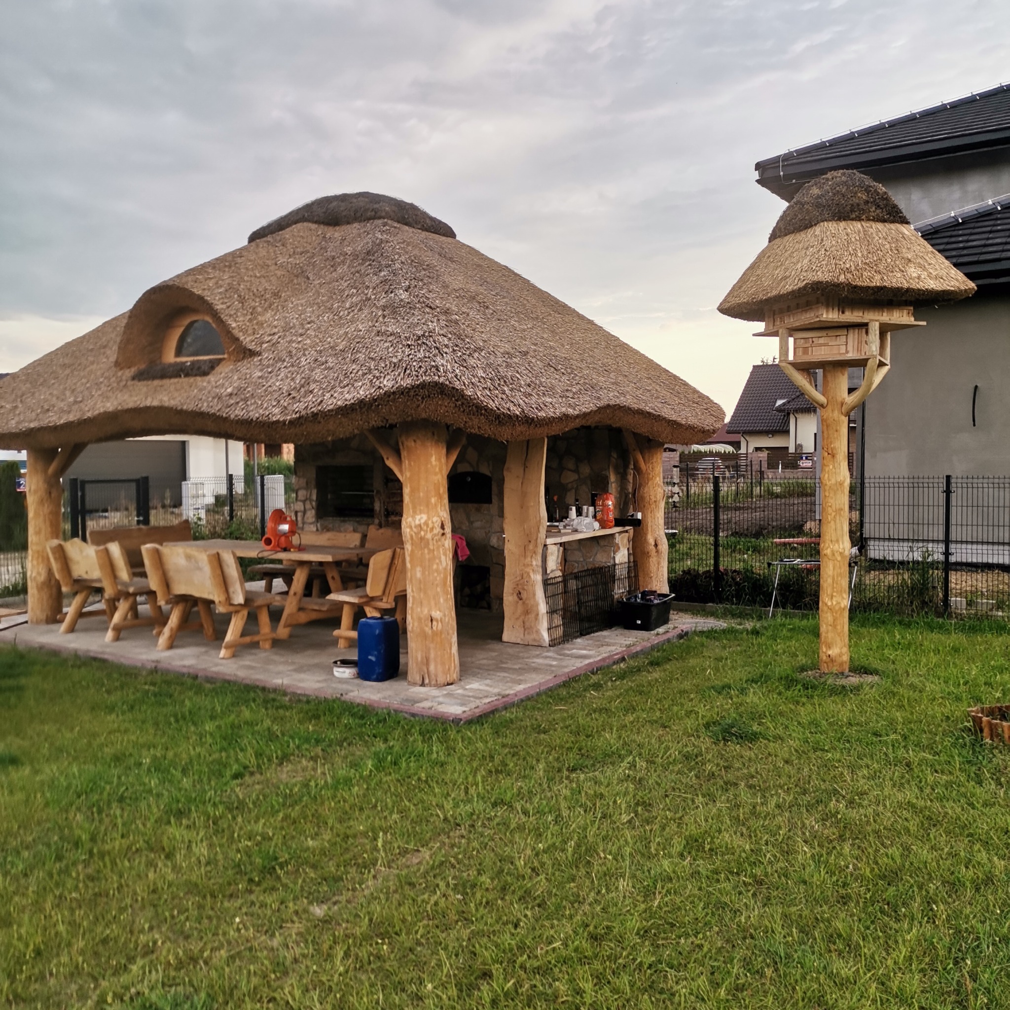 Ogród z altaną krytą strzechą, drewnianym stołem, ławami i grillem murowanym, obok karmnik dla ptaków również kryty strzechą.