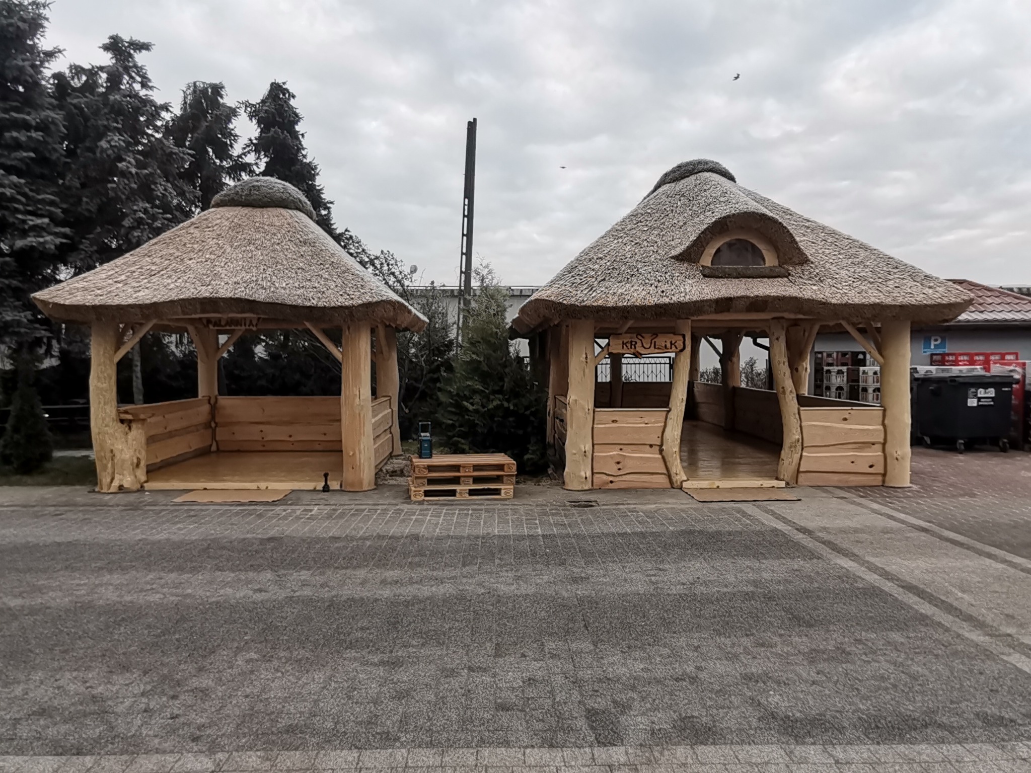 Dwie drewniane konstrukcje z trzcinowymi dachami, jedna z owalnym oknem, druga z paletą przed wejściem, na tle szarej kostki brukowej i pochmurnego nieba.