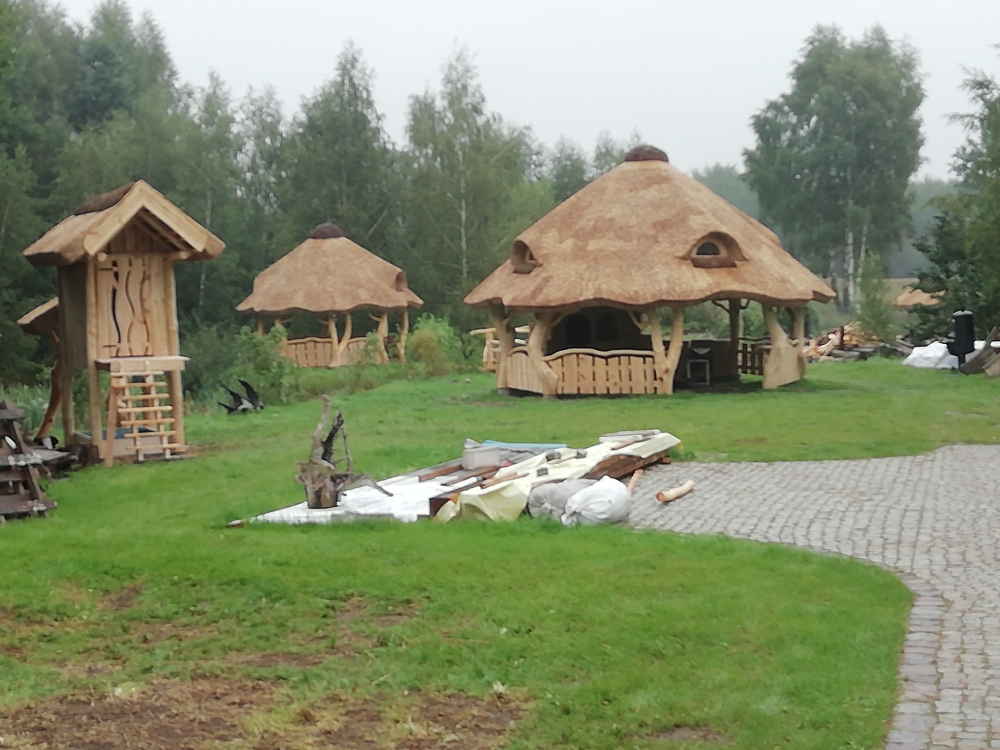 Trzy konstrukcje z krytymi strzechą dachami: mała wieża widokowa, altana z ażurową balustradą i altana z okienkami, na trawniku widoczne materiały budowlane.