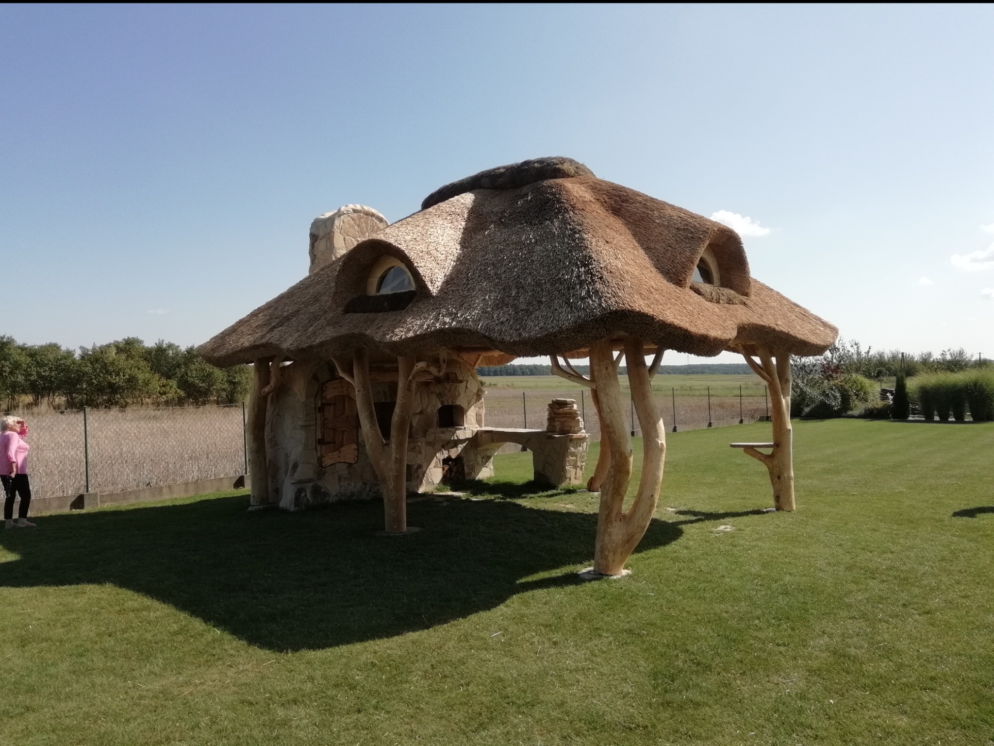 Oryginalna altana z krytym strzechą dachem, wsparta na pniach drzew, z kamiennym piecem i kominem, z widocznym fragmentem krajobrazu i osobą w oddali.