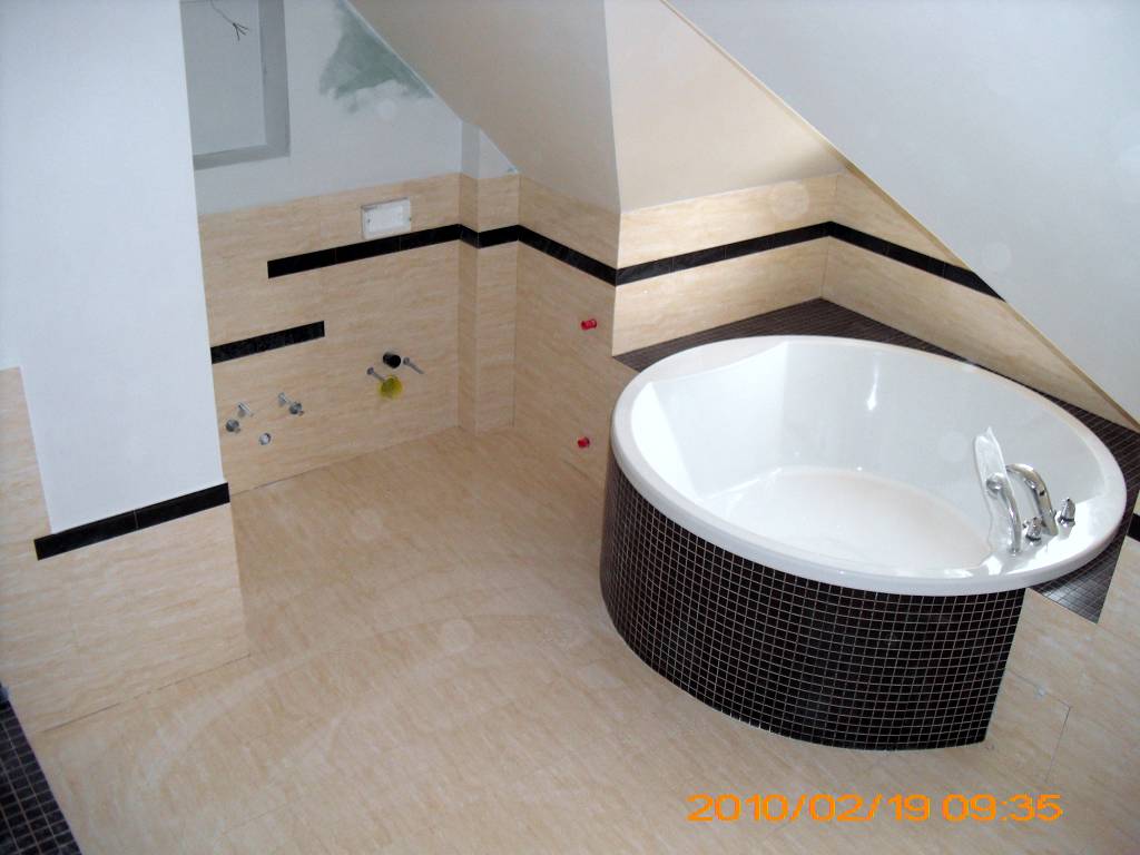 Niedokończona łazienka na poddaszu z wanną obudowaną czarną mozaiką, beżowe płytki na ścianach i podłodze, widoczne wystające rury i czerwone kliny dystansowe do płytek.