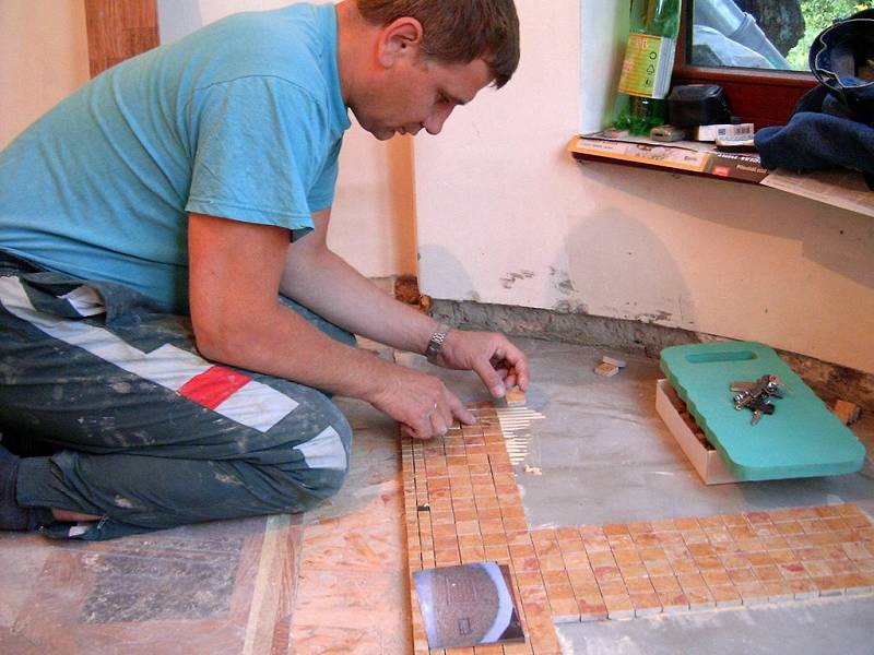 Mężczyzna w kombinezonie układa mozaikę z małych, brązowych płytek na podłodze w pomieszczeniu, obok widoczna zielona podkładka z kluczami i zdjęcie.