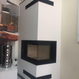 kominkiwojnoto - Biały, narożny wkład kominkowy z czarnymi akcentami, widoczne akcesoria kominkowe i aranżacja wnętrza w tle.