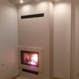 kominkiwojnoto - Nowoczesny, wnękowy palenisko z płomieniem, obudowane beżowym kamieniem, wbudowane w ścianę z minimalistycznym, czarnym grillem wentylacyjnym u góry.