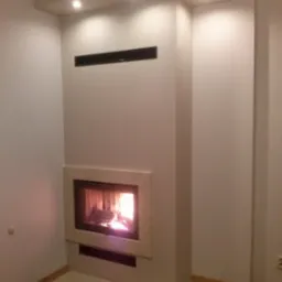 Nowoczesny, wnękowy palenisko z płomieniem, obudowane beżowym kamieniem, wbudowane w ścianę z minimalistycznym, czarnym grillem wentylacyjnym u góry.