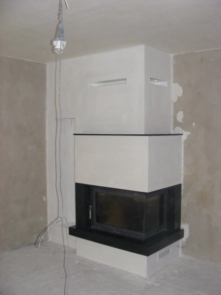 Nowoczesny, narożny wkład kominkowy z czarną obudową, widoczny w surowym wnętrzu w trakcie wykończenia, z wiszącą żarówką i odsłoniętymi przewodami.