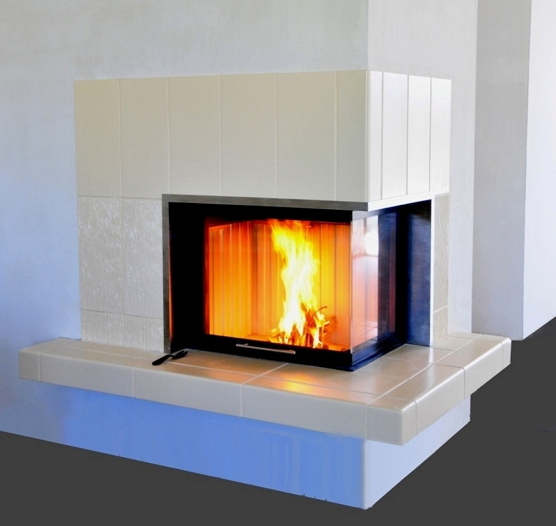 Narożny wkład kominkowy z szybą, obudowany białymi płytkami ceramicznymi, z palącym się wewnątrz ogniem, widoczna czarna rączka do otwierania drzwiczek.