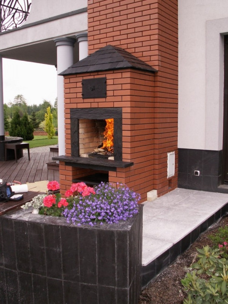 Murowany, zewnętrzny piec z paleniskiem, obudowany czerwoną cegłą, z czarnym dachem i zamknięciem, płomienie wewnątrz, obok kwitnące kwiaty w donicy.
