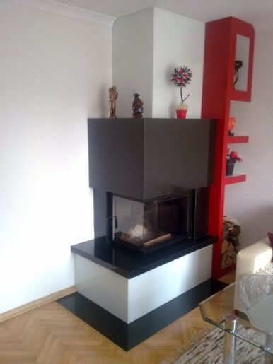 Nowoczesny kominek narożny z czarnym obramowaniem i białym dolnym panelem, na górze dekoracje: figurka i sztuczny kwiat, obok czerwony regał.