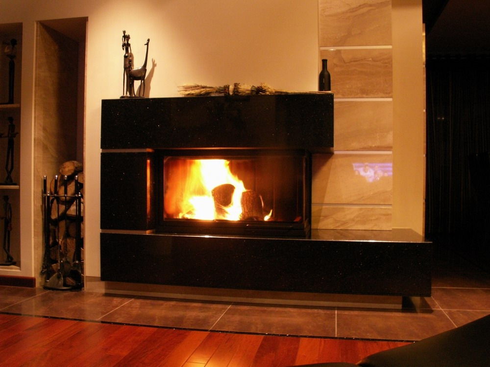 Nowoczesny, czarny kominek z paleniskiem, ozdobiony afrykańskimi figurkami i butelką, z drewnem ułożonym obok i palącym się ogniem wewnątrz.