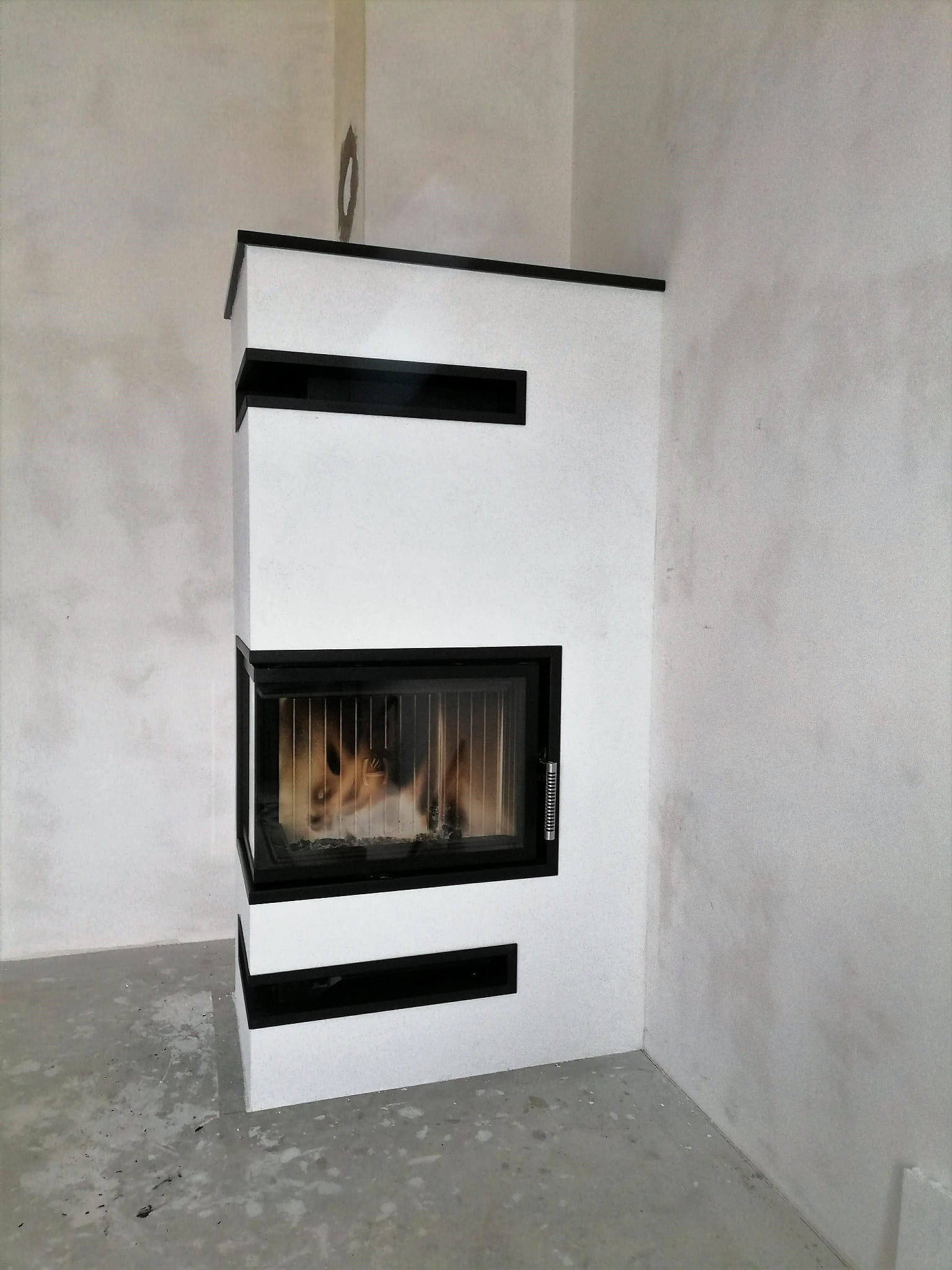 Nowoczesny, narożny piec kaflowy z czarnymi akcentami, płomienie widoczne przez szybę, surowe wykończenie ścian w tle.