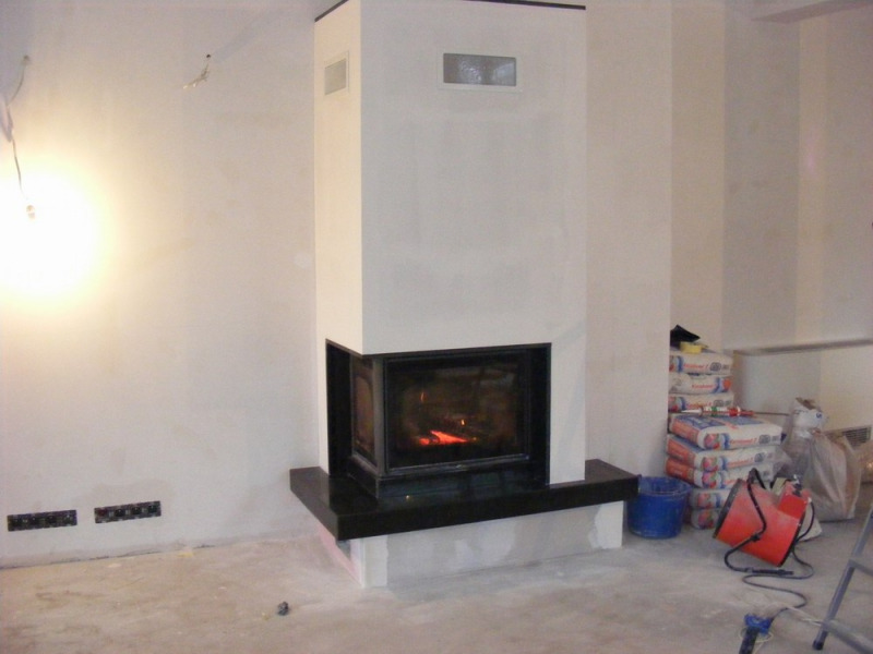 Narożny kominek z czarnym obramowaniem i marmurowym blatem w surowym wnętrzu w trakcie wykańczania, widoczne worki z zaprawą, grzejnik i drabina.