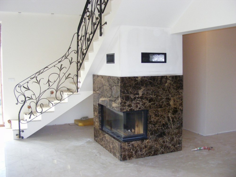 Narożny wkład kominkowy obudowany brązowym marmurem, widoczny pod schodami z ozdobną, kutą balustradą w jasnym wnętrzu, w trakcie wykańczania.