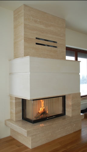 Nowoczesny, narożny kominek z obudową z trawertynu i białym elementem nad paleniskiem, z widocznym płomieniem i drewnem wewnątrz.