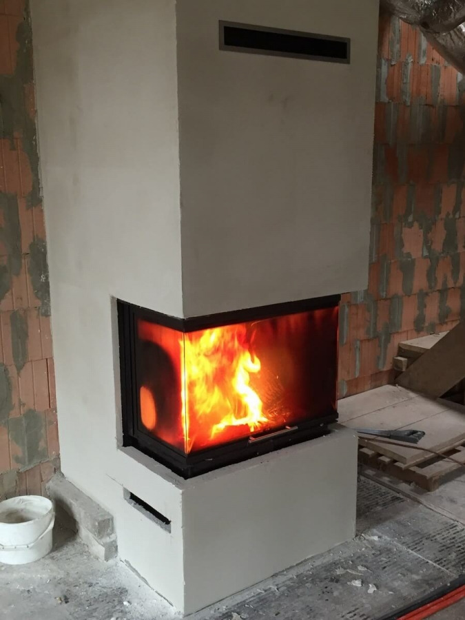 Nowoczesny, narożny wkład kominkowy z płomieniami, obudowany surowym, otynkowanym korpusem w trakcie budowy, widoczne cegły w tle i wiadro z zaprawą.