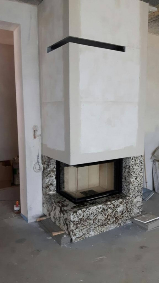 Wnętrze w trakcie budowy z nowoczesnym, narożnym kominkiem obłożonym płytami imitującymi granit, z widocznymi elementami konstrukcyjnymi i niedokończonymi ścianami.