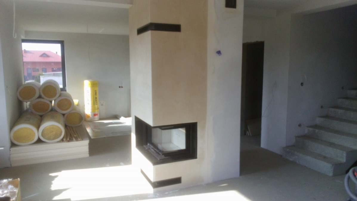 Wnętrze w trakcie wykańczania z nowoczesnym narożnym wkładem kominkowym z czarną ramą, widoczne rolki izolacji w tle przy oknie.