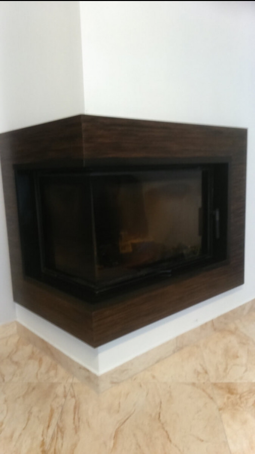 Wbudowany, narożny wkład kominkowy z ciemną, drewnopodobną obudową, widoczny z perspektywy pod kątem, z marmurkową posadzką w tle.