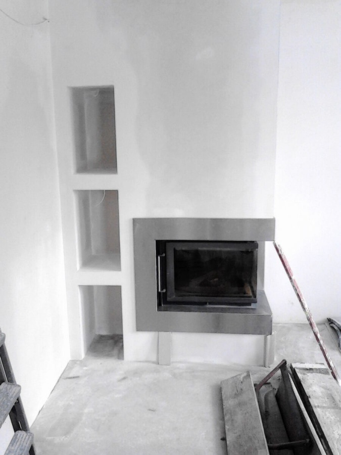 Surowy stan montażu: nowoczesny wkład kominkowy z metalową obudową, wmurowany w białą ścianę z trzema niszami po lewej stronie, widoczne narzędzia i drabina w tle.