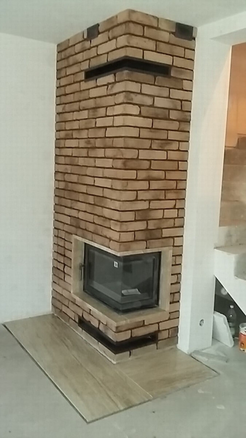 Murowany, narożny piec z czarnym wkładem kominkowym, wykończony cegłą klinkierową w odcieniach beżu i brązu, z widoczną częścią posadzki wyłożonej płytkami imitującymi drewno.