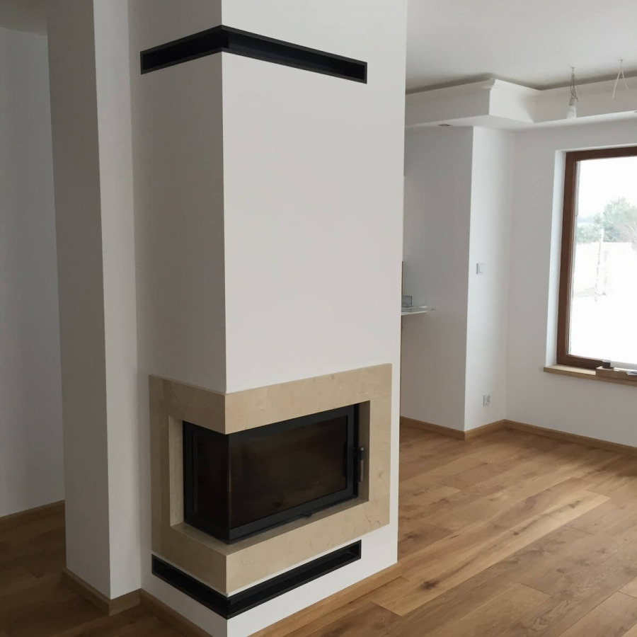 Nowoczesny narożny wkład kominkowy z obudową z jasnego kamienia, z czarnymi akcentami w górnej i dolnej części, na tle jasnego wnętrza z drewnianą podłogą.